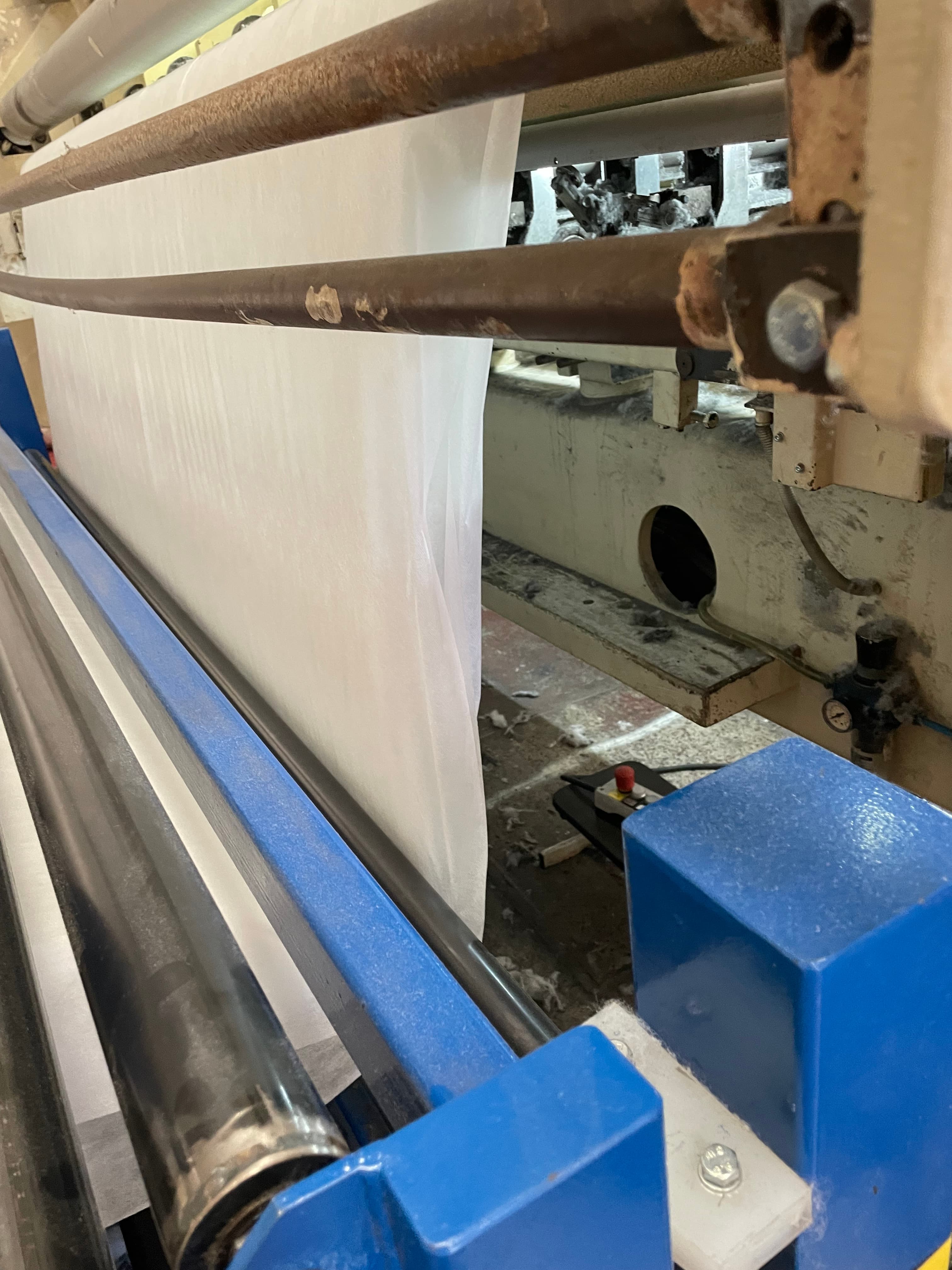 Manufacturing floor — blue-framed machine with worn brown rollers, white fabric running through, fiber dust on floor